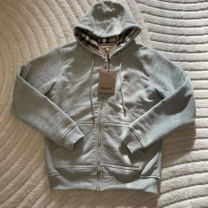 Burberry Hoodie - Snygg grå hoodie från Burberry med dragkedja och klassiskt rutigt foder i huvan. Perfekt för en stilren och avslappnad look.