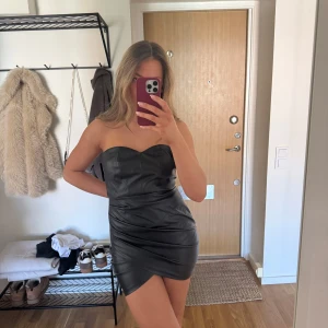 Svart axelbandslös klänning i skinnimitation - Säljer en svart axelbandslös klänning i skinnimitation med tight passform och kort längd. Klänningen har en stilren och modern look, perfekt för dig som vill sticka ut. Snyggt formad upptill och markerar midjan.
