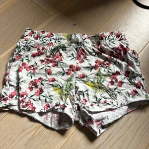 Blommiga shorts med fågelmotiv från Basic Needs är för barn  - Säljer ett par vita shorts med elastisk midja från Basic Needs. Shortsen har ett färgglatt blommönster i rött och grönt samt stora gula fåglar. Perfekta för varma dagar och riktigt sköna att ha på sig. Är i storleken 146/cr 11 år