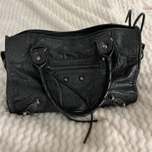 Svart handväska med dragkedja och detaljer - Säljer en svart handväska med klassisk design och silverfärgade metalldetaljer. Väskan har både handtag och dragkedja upptill samt en mindre ficka med dragkedja framtill. Materialet är syntet med en lätt glansig yta och flätade detaljer på handtagen.