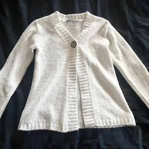 Vit stickad kofta från Kids by Lindex - Mysig vit stickad kofta från Kids by Lindex i barnstorlek 110/116. Koftan har lång ärm, bred ribbad kant och stängs med en stor knapp framtill. Perfekt för lager-på-lager under kyliga dagar. Säljs för 80kr+frakt.