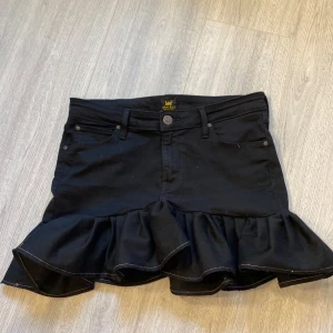 Svart jeanskjol med volang - Supersnygg svart kortkjol med volang nertill. Klassisk jeansmodell med fem fickor och knappstängning framtill. Perfekt för dig som vill ha en kjol med lite extra detaljer. Har sytt den själv utav ett par Lee jeans, skulle säga s/m