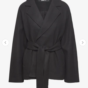 Svart kappa  - Stilren svart kappa från Bikbok med knytbälte i midjan och klassisk krage. Jackan har lång ärm och en rak, något oversized passform. Perfekt för kyligare dagar när du vill vara både snygg och bekväm.