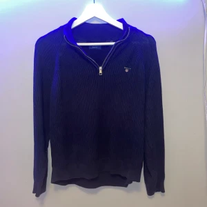 Mörkblå stickad tröja med dragkedja från Gant - Säljer en mörkblå stickad tröja från Gant med halv dragkedja och liten broderad logga på bröstet. Tröjan har lång ärm och ribbade muddar vid ärmslut och nederkant. Perfekt för lager-på-lager-stil.