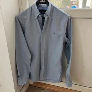Blå skjorta från Ralph Lauren - Klassisk lblå skjorta från Ralph Lauren i slim fit-modell. Skjortan har knappar framtill, button-down krage och den ikoniska broderade loggan på bröstet. Perfekt för en stilren look. Pris kan diskuteras ny pris 1500 kr!