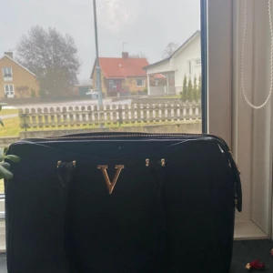 Svart handväska med gulddetaljer - Säljer en svart handväska från Valentino med guldfärgad V-detalj framtill och dragkedja upptill. Väskan har två handtag och flera fack inuti, inklusive innerficka med dragkedja. Perfekt för att bära dina viktigaste saker.Frakt tillkommer