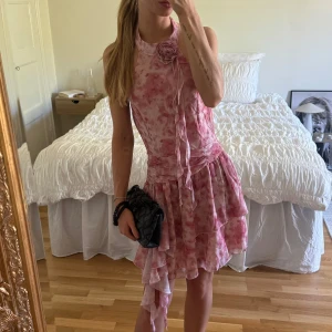 Rosa volangklänning med blommönster - Rosa klänning med volanger och blommigt mönster. Klänningen har ärmlös design, öppen rygg och asymmetrisk nederdel. Perfekt för sommaren!