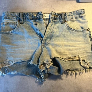 Ljusblå slitna jeansshorts med fransar - Säljer ett par ljusblå jeansshorts med råa kanter och slitna detaljer framtill och baktill. Shortsen har klassisk femficksdesign, knappar i gylfen och fransar längs bensluten. Perfekta för en avslappnad stil.