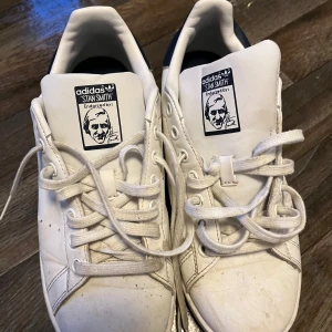Vita Adidas Stan Smith sneakers - Klassiska vita Adidas Stan Smith sneakers med blå detalj på hälen och perforerade ränder på sidorna. Skorna har snörning och platt sula, samt Stan Smith-logga på plösen. Perfekta för en stilren och tidlös look.