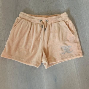 Peachfärgade frottéshorts från Juicy Couture - Snygga peachfärgade shorts från Juicy Couture i mjuk frotté med dragsko i midjan och fickor fram. På vänster ben finns en glittrig JC-logga. Perfekta för sommardagar eller chilla hemma.