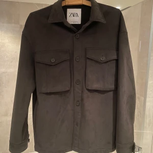 Olivgrön Mocka Jacka - Stilren olivgrön från Zara med klassisk krage och två stora bröstfickor med lock och knappar. Jackan har knäppning framtill och långa ärmar med knapp vid ärmslut. Perfekt för lager-på-lager under sommar och vår.