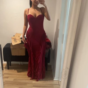 Vinröd långklänning med volangdetaljer - Säljer en vinröd långklänning med smala axelband och volangdetaljer längs sidorna. Klänningen har en figurnära passform, öppen rygg och djup urringning framtill. Perfekt för speciella tillfällen.