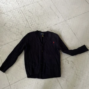 Mörkblå kabelstickad kofta från Polo Ralph Lauren - Mörkblå kabelstickad kofta från Polo Ralph Lauren med knappar framtill och klassisk röd logga på bröstet. Långärmad modell med ribbade muddar och v-ringad hals. Perfekt för lager-på-lager och stilren look. En knapp fattas