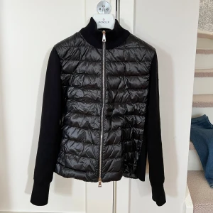 Svart cardigan från Moncler - Säljer en svart padded wool zip-up från Moncler i svart, den är i storlek xs. Jackan har hög krage och dragkedja framtill. Klassisk Moncler-logga på ena ärmen. Köpt på NK förra våren, nypris 10299kr. Den är använd ett fåtal gånger och är som ny. Inget att anmärka på. Kvitto finns. 