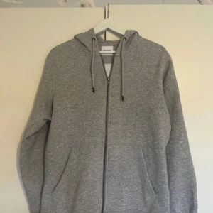 Grå hoodie med dragkedja från Jack and Jones  - En klassisk grå hoodie från Weekday med dragkedja framtill och två fickor. Tröjan har huva med dragsko och är tillverkad i mjukt material. Perfekt för en avslappnad stil.