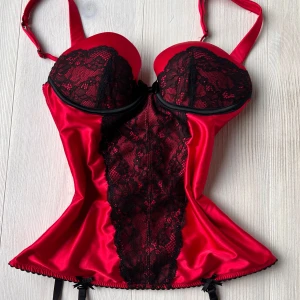 Röd och svart korsettopp med spets - Snygg röd korsettopp med svarta spetsdetaljer och rosett framtill. Toppen har vadderade kupor och justerbara axelband. Perfekt för att skapa en statement-look.