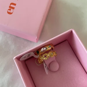 Guldfärgade ringar med stenar - Aldrig använda! Två guldfärgade ringar med dekorativa stenar i grönt, rosa och vitt. Ringarna har ett unikt mönster med infattade stenar runt om och levereras i en rosa ask. Perfekt för att ge din look lite extra färg och glans. Går även köpa en av ringarna för 180kr.