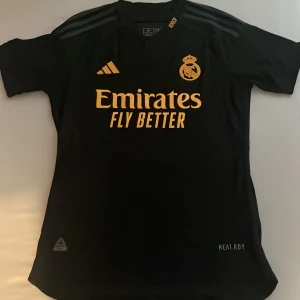Real Madrid svart fotbollströja Bellingham 5 - Säljer en svart Real Madrid fotbollströja från Adidas med gula detaljer och tryck. Tröjan har korta ärmar, klubbmärke och sponsortryck på framsidan samt Bellingham 5 på ryggen. Tillverkad i lätt funktionsmaterial. Spelarversion, äkta och nyskick.