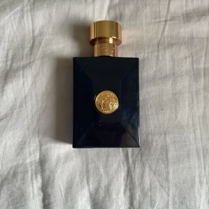Versace Dylan Blue Parfym - En lyxig herrparfym från Versace i en elegant mörkblå glasflaska med guldigt lock och Versaces ikoniska Medusa-emblem framtill. Flaskan har en stilren, fyrkantig form och utstrålar exklusivitet. Den luktar citrus och sommar.