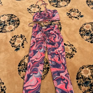 Färgglada byxor och topp med snörning - Säljer ett set med byxor och matchande topp i färgglatt mönster med rosa, lila och blå toner. Byxorna har normal passform och snörning framtill. Topp med smala axelband och snörning. Perfekt för dig som gillar unika plagg!
