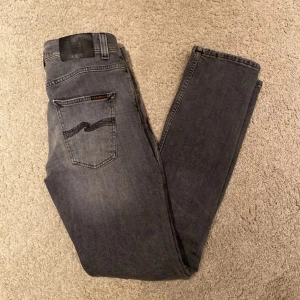 Grå jeansbyxor från nudie - Säljer ett par grå jeansbyxor från nudie Jeans med klassisk femficksdesign och diskret broderad detalj på bakfickan. Byxorna har normal passform och raka ben, perfekta till vardagsstilen.