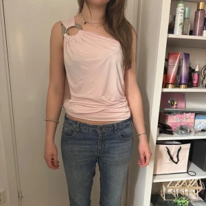Ljusrosa oneshoulder topp med ringdetalj - Säljer en ljusrosa oneshoulder topp med en snygg ringdetalj på axeln. Toppen har en draperad passform och öppen rygg, vilket ger en cool och trendig look. Perfekt för dig som vill sticka ut lite extra.