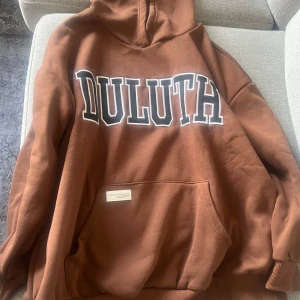 Brun hoodie med DULUTH-tryck - Säljer en brun hoodie med stor vit och svart DULUTH-text på bröstet. Tröjan har huva och en stor magficka framtill. Perfekt för en avslappnad och cool stil.