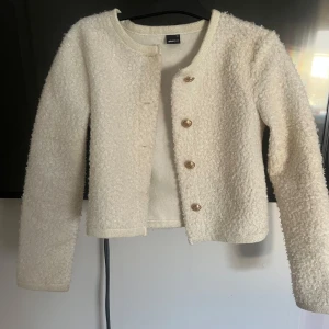 Vit teddyjacka från Gina Tricot - Säljer en söt vit teddyjacka från Gina Tricot i storlek XS. Jackan har rund halsringning, guldiga knappar framtill och är croppad i modellen. Perfekt för att ge en mysig och trendig look.