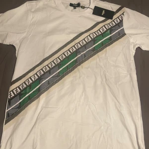 Vit t-shirt med diagonal Fendi-logga - Säljer en vit t-shirt från Fendi med en diagonal rand över bröstet i svart, grått, grönt och beige. Randen har Fendi-loggor och ger en unik look. Perfekt för dig som gillar designerplagg. Aldrig använt.