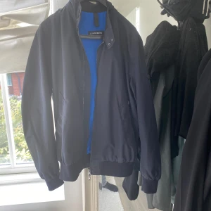 Mörkblåjacka från J.Lindeberg - Snygg mörkblåjacka från J.Lindeberg med dragkedja framtill och ribbade muddar vid ärmslut och midja. Jackan har en klassisk passform och diskret logotyp på ärmen. Perfekt för en stilren look. Endast använd ett fåtal gånger så jackan är som ny. Storlek: S Skick:9/10