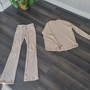 Beige ribbade byxor och tröja från Stronger - Säljer ett par beige ribbade byxor från Stronger med normal passform och dragsko i midjan. Byxorna har vida ben och är tillverkade i mjukt material som passar perfekt för en avslappnad stil.