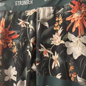 Blommiga tights från Stronger - Snygga tights med blommigt mönster i orange, vitt och grönt på mörk botten. Hög midja och elastisk linning med Stronger-logga. Perfekta för träning eller en färgglad vardagslook.