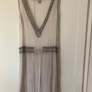 Vit klänning med pärldetaljer - Säljer en elegant vit klänning från Noa Noa med v-ringning och pärlbroderade detaljer längs halsringning, midja och i kjolen. Klänningen är ärmlös och det följer med en vit underklänning. Perfekt för sommarens speciella tillfällen. Storlek M motsvarar 40. Väldigt sparsamt använd. 