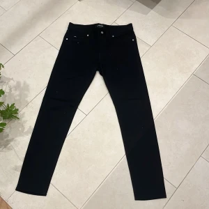 Svarta jeans från J.Lindeberg - Svarta jeans från J.Lindeberg med klassisk femficksdesign och raka ben. Byxorna har diskret logga på myntfickan och stängs med dragkedja och knapp. Perfekta för en stilren och enkel look. PRIS KAN DISKUTERAS!