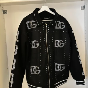 Dolce & Gabbana jacka - Säljer en svart jacka från Dolce & Gabbana med stora DG-loggor och siffror i strass över hela jackan. Jackan har dragkedja framtill, lång ärm och vita ränder vid muddar och nederkant. 100% bomull och tillverkad i Italien.
