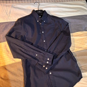 East West bomulls skjorta  - Säljer en klassisk och snygg skjorta från East West i mörkblå färg. Modellen har button-down-krage och vita knappar, vilket ger en tidlös och välklädd look. Passform: Slimfit 🙌⭐️