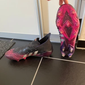 Adidas Predator fotbollsskor i svart och rosa - Säljer ett par Adidas Predator fotbollsskor i svart och rosa. Skorna har en strumpliknande passform och mönstrad ovansida för extra grepp på bollen. Finns hål för snören men inge snören följer med. En liten skada finns på höger skon men inget jätte allvarligt (se sista bilden). Perfekta för dig som vill sticka ut på planen!