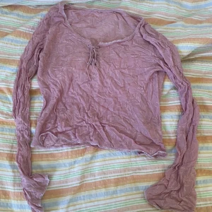 Ljuslila spetsig långärmad topp - Säljer en ljuslila långärmad topp med spetsdetaljer och broderat bladmönster. Toppen har knyt framtill vid urringningen och är tillverkad i ett tunt, genomskinligt material. Perfekt för lager-på-lager och en söt detalj till outfiten.