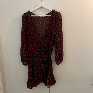 Svart omlottklänning med röda hjärtan från KILIBBI - Supersöt svart omlottklänning från KILIBBI med röda hjärtan, långarmar och volangkant nertill. Klänningen har knyt i midjan och är kort i modellen. 