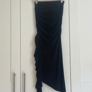 Svart midikjol med volang från Gina Tricot - Säljer en svart midikjol från Gina Tricot med snygg volangdetalj längs sidan. Kjolen har en draperad effekt och asymmetrisk nederkant som ger en cool och trendig look. Perfekt att styla med en enkel topp och boots.