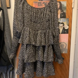 Svart blommig volangklänning - Supersöt svart klänning med vitt blommönster och flera volanglager. Långärmad modell med smockad överdel och rund halsringning. Perfekt för dig som gillar romantisk och trendig stil.