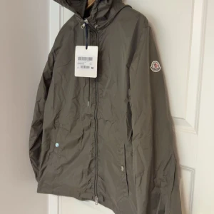 Olivgrön vindjacka från Moncler - Snygg olivgrön vindjacka från Moncler med huva som har röd, vit och blå rand. Jackan har dragkedja framtill, två fickor och Moncler-logga på ärmen. Perfekt för blåsiga dagar och enkel att matcha med olika outfits.