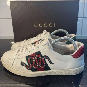 Gucci Ace sneakers med ormmotiv - Säljer ett par vita Gucci Ace sneakers med klassiskt ormmotiv broderat på sidan och röda samt gröna detaljer på hälen. Skorna har snörning och platt sula. Perfekta för dig som vill sticka ut med en ikonisk design. Storlek uk 7 eu 42