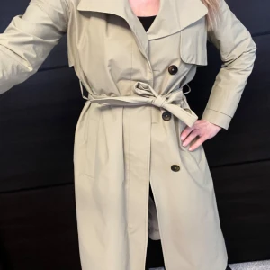 Beige trenchcoat från Tiger - Snygg beige trenchcoat från Tiger med knytskärp i midjan och stora knappar framtill. Jackan har klassisk krage och slät, glansig insida. Perfekt för dig som vill ha en stilren och tidlös look.