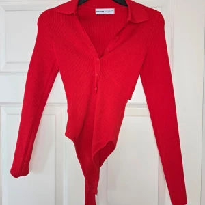 Röd ribbad bodysuit med krage från Bershka - Säljer en röd ribbad bodysuit från Bershka med klassisk krage och knappar framtill. Bodyn har lång ärm och är figurnära i modellen. Perfekt att styla med jeans eller kjol för en trendig look. Helt ny/aldrig använd.