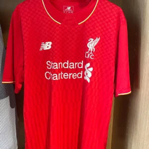 Liverpool FC röd fotbollströja från New Balance 15/16 - Säljer en röd Liverpool FC fotbollströja från New Balance med klubbmärke och sponsortryck på bröstet. Tröjan är köpt för 7 år sedan och har bara legat i garderoben då den är för stor för mig. Den är äkta och köpt i en riktig butik. Detta är en väldigt unik tröja som är svår att få tag på, speciellt för den är äkta och bra kvalité!
