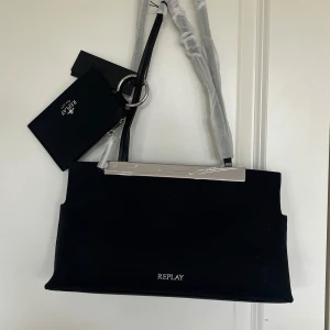 Svart axelväska från Replay med silverdetaljer - Snygg svart axelväska från Replay med stilrent silverfärgat metallhandtag och tillhörande svart plånbok med krokodilmönster. Väskan har minimalistisk design och logga framtill. Perfekt för dig som gillar stilrena accessoarer.