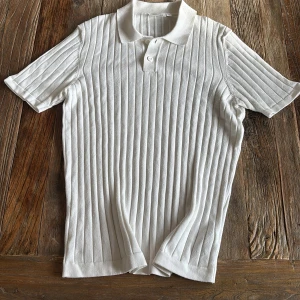 Vit ribbad pikétröja från Kappahl - Säljer en stilren vit pikétröja från Kappahl i ribbat material. Tröjan har korta ärmar, klassisk krage och knäppning framtill. Tillverkad av viskos och polyester för en mjuk och bekväm känsla. Perfekt för en fräsch och enkel look. Använd 1-2 ggr