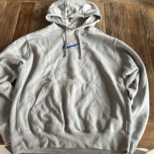 Grå hoodie från Nike med blå AIR-logga - Säljer en grå hoodie från Nike i storlek M. Tröjan har en stor blå AIR-logga med swoosh på ryggen och mindre logga framtill. Klassisk känguruficka och justerbar huva med dragsko. Tillverkad i mjuk bomull för extra komfort. Använd 1-2 ggr