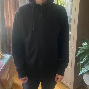 Svart hoodie med goggles från C.P. Company - Säljer en svart hoodie från C.P. Company med deras ikoniska goggles i huvan. Jackan har dragkedja framtill, justerbar huva med snörning och lång ärm. En knapp har lossnat samt har ett av glasögonen lossnat och går ej att sätta tillbaka (Se bild). Borde gå att laga hos skräddare. Storlek Medium och jag är 176cm